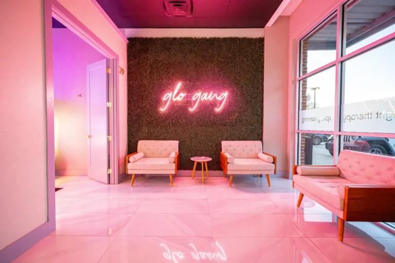 A foyer with two chairs, small table, and a sofa at a tanning salon with a pink "glo gang" illuminated sign.