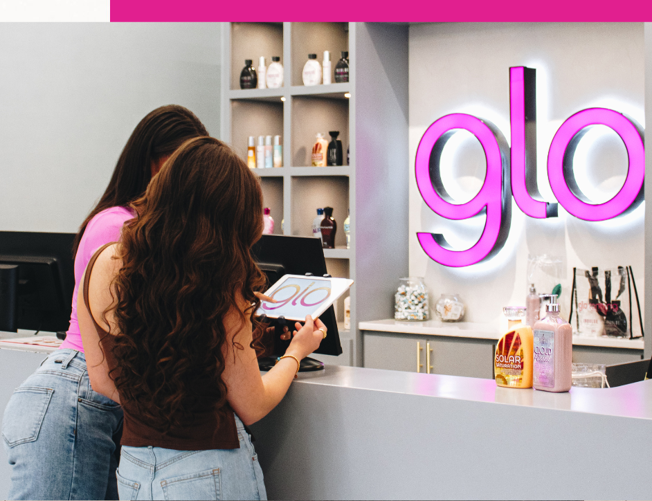 Two women looking at a tablet at a Glo Tanning salon reception desk.