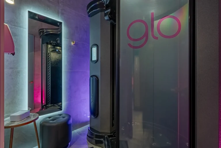 Vertical tanning booth labeled glo next to a glowing mirror, gray stool, and small round wooden table.