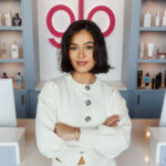 Woman in white outfit smiling with crossed arms at a tanning salon reception desk, glo sign and beauty products behind her.